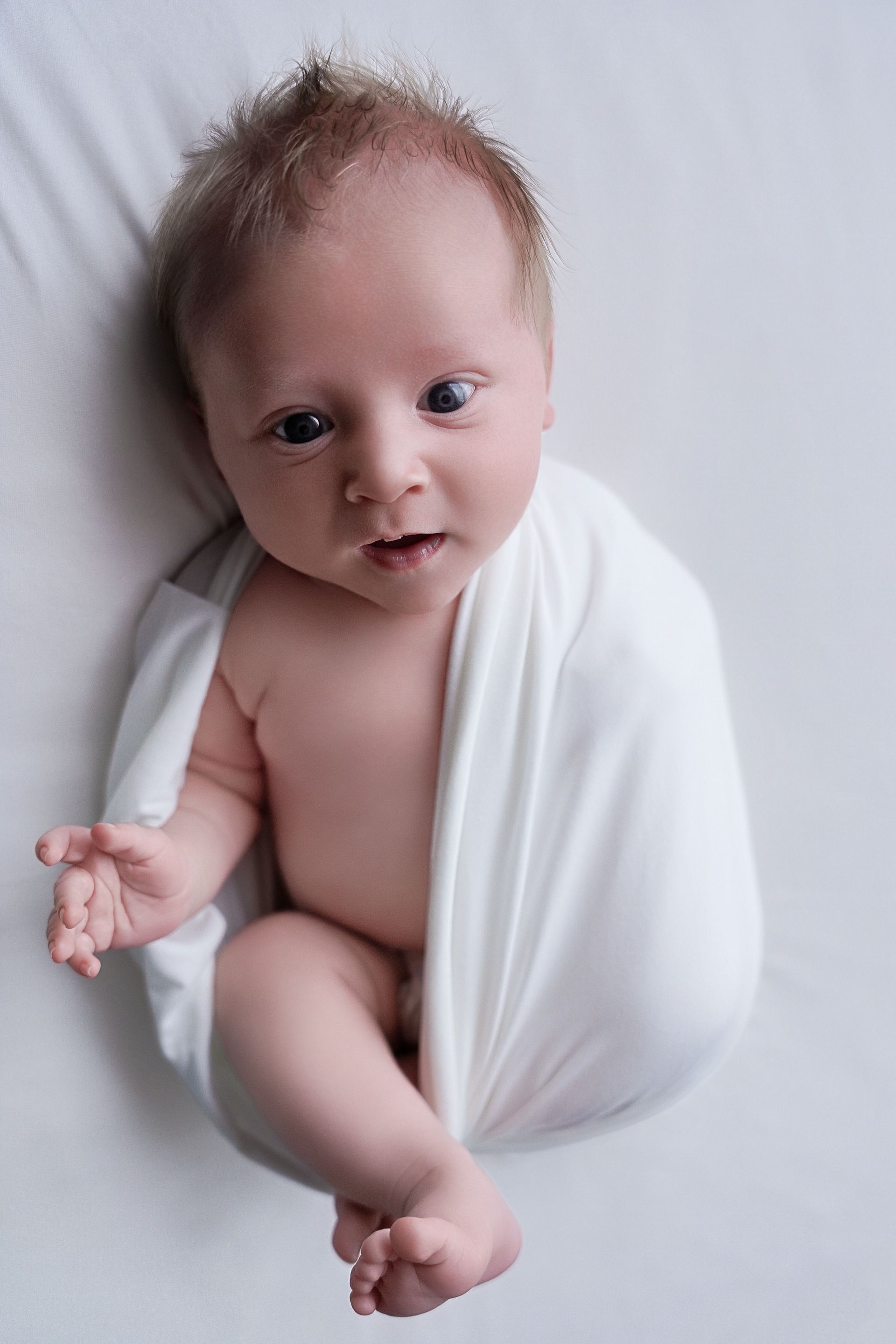 De baby, gewikkeld in een witte doek, ligt op een zachte witte deken en kijkt omhoog.