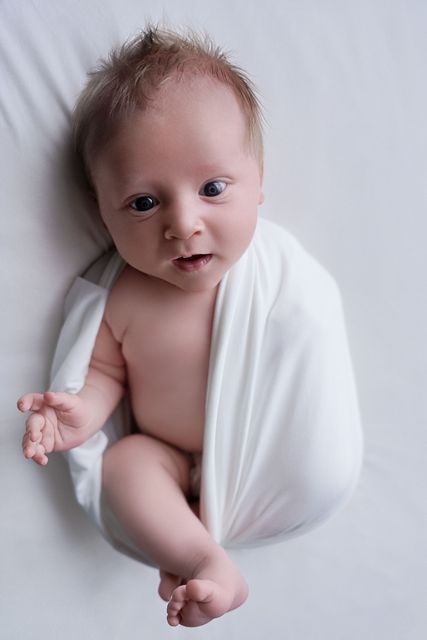 De baby, gewikkeld in een witte doek, ligt op een zachte witte deken en kijkt omhoog.
