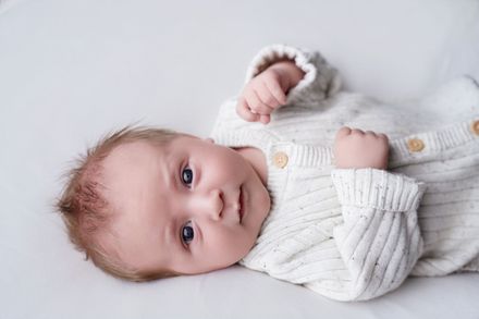 De baby ligt op een witte deken, draagt een crèmekleurig gebreid pakje en kijkt naar de camera.