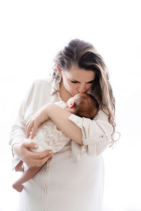 Vrouw in een witte jurk houdt een slapende baby teder vast en kust hem, tegen een helderwitte achtergrond.