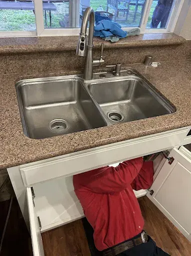Person reaching into under-sink cabinet beneath a double kitchen sink with a window above the counter.