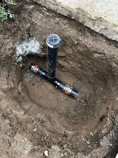 Excavated trench showing a black vertical pipe and horizontal pipe connections in dirt.