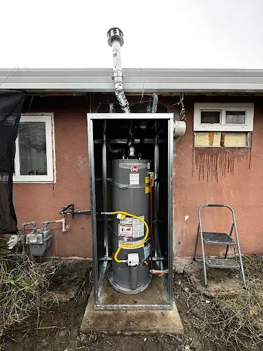 Water heater in an open metal enclosure beside a house, with a ladder and window nearby.