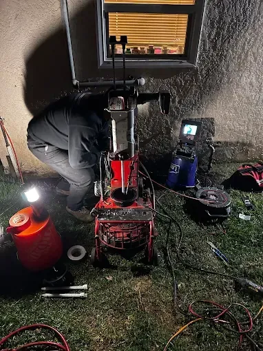 Technician working on a red machine outdoors at night, lit by work lights beside a window.