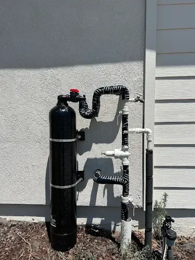 Black water softener tank and exposed pipes beside a house foundation.