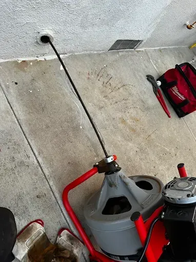Red plumbing snake with black cable feeding into a wall vent on a concrete floor, next to a red tool bag