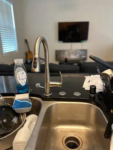 Kitchen sink with faucet, dish soap, and cleaning brush; living room TV in the background