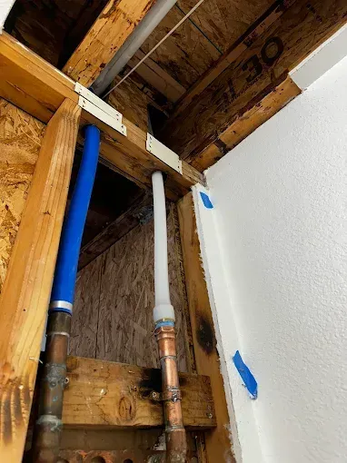 Open ceiling with exposed blue and white pipes beside a white wall and wooden framing