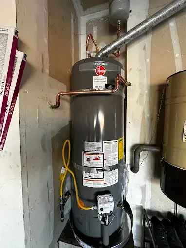 Water heater in a utility closet with exposed pipes and a yellow hose.