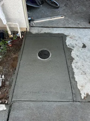 Concrete utility pad with a circular drain opening beside a building entrance.