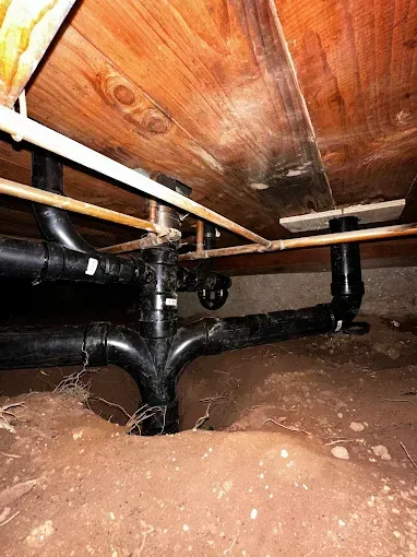 Exposed black pipes and a dirt floor beneath a wooden structure, showing a crawlspace or underfloor area