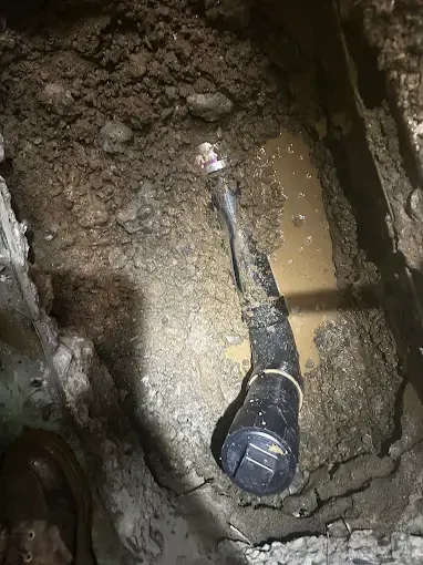 A black pipe with a coupling lies in a muddy trench with standing water and exposed dirt sides.