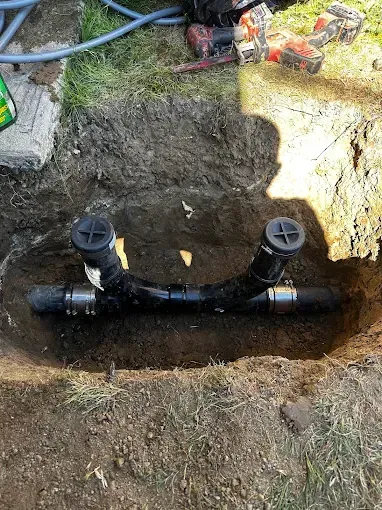 Black plumbing pipe and T-joint installed in a dirt trench outdoors