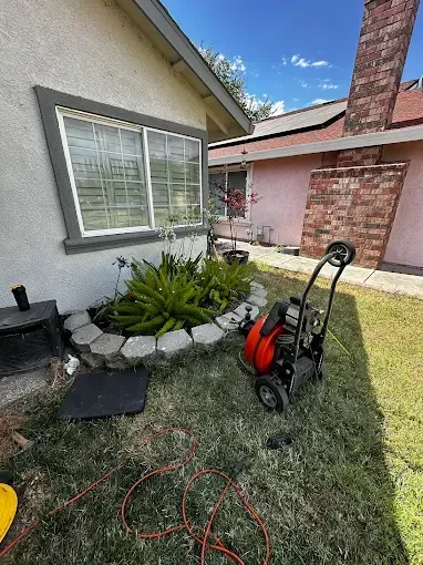 Side yard with hose reel, coiled red cord, stone-bordered plants, and a house wall with a window.