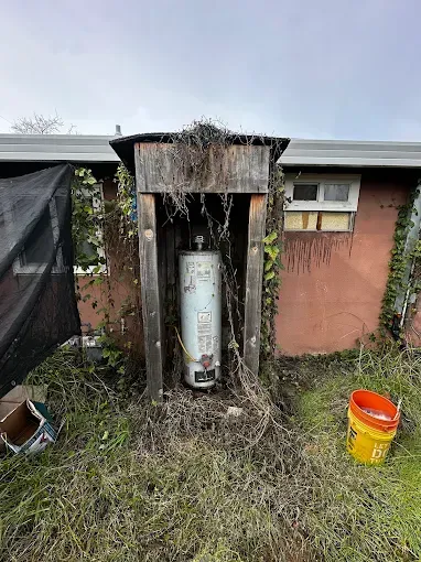Outdoor water heater in a weathered shed beside a house, with a yellow bucket and overgrown grass.