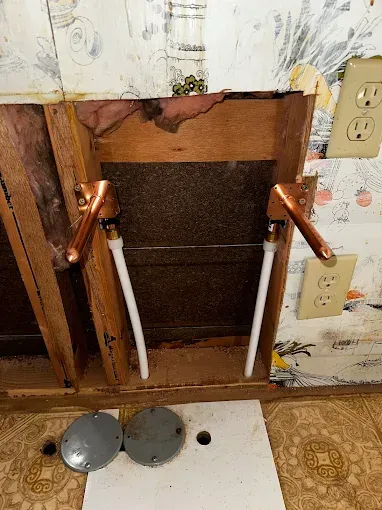Open wall cavity with exposed plumbing pipes and a wall outlet beside damaged drywall.