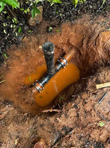 Orange underground pipe exposed in a muddy trench, with a black vent pipe attached.