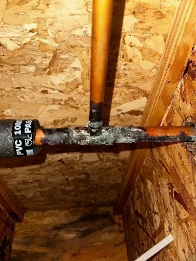 Corroded black and copper pipes in a plywood wall cavity, with white PVC pipe label visible