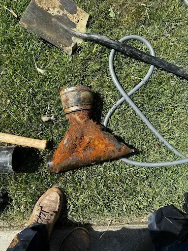 Rusty triangular metal tool attachment on grass, with a shovel and gray hose nearby.