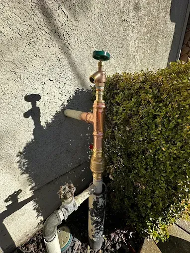 Outdoor water shutoff valve and pipes beside a stucco wall and hedge.