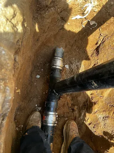 Feet beside an exposed black pipe and metal fitting in a dirt trench.