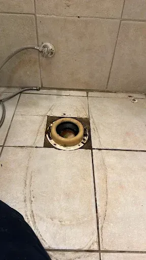 Bathroom floor with an exposed toilet drain hole and a loose hose against beige tiles.