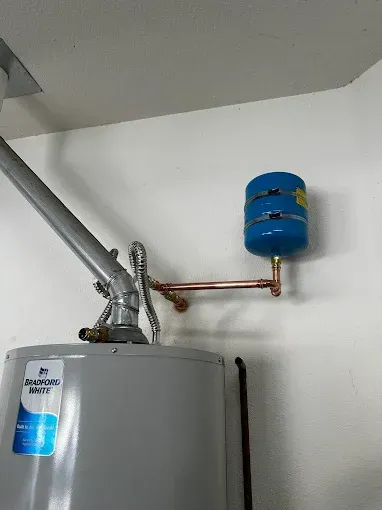 Water heater with metal vent and blue expansion tank on a white wall.
