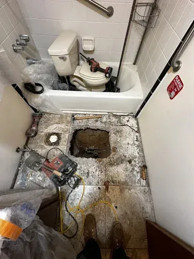 Small bathroom with damaged floor, exposed hole, tools, and a toilet in the back corner.