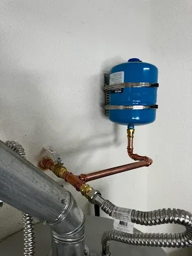 Blue expansion tank and copper plumbing connected to metal ducts on a white wall.