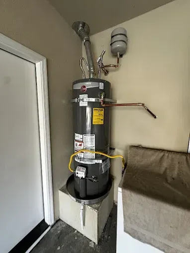 Gas water heater in a utility closet beside a white door, with vent pipe and wall-mounted vent fan.