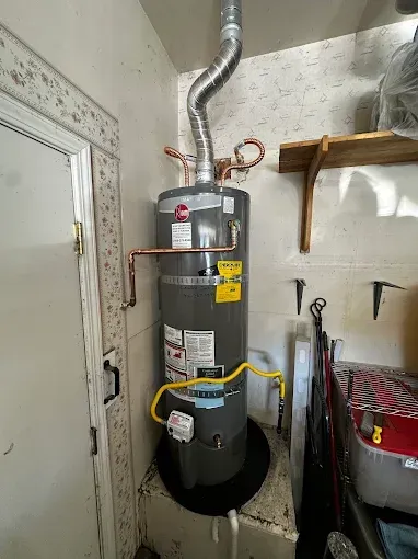Water heater in a small utility closet with exposed pipes, vents, and a yellow gas line.