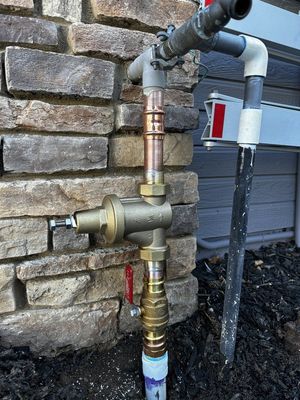 Outdoor plumbing pipes and valves mounted on a stone wall beside running water.