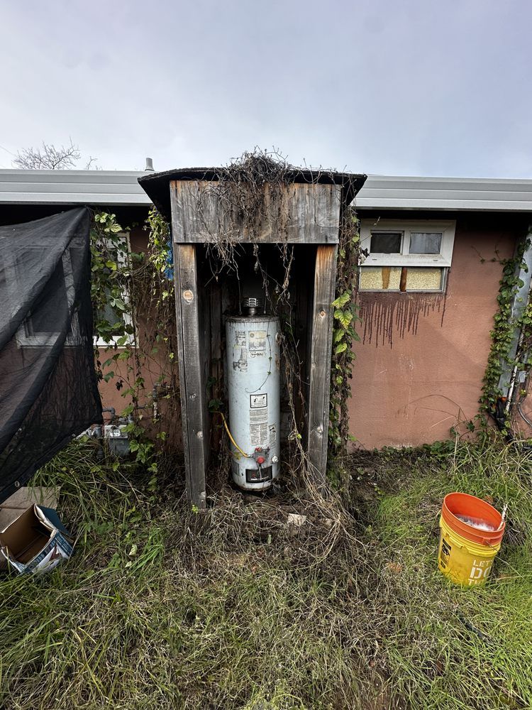 Weathered outdoor water heater with a black tarp and orange bucket beside a house wall.