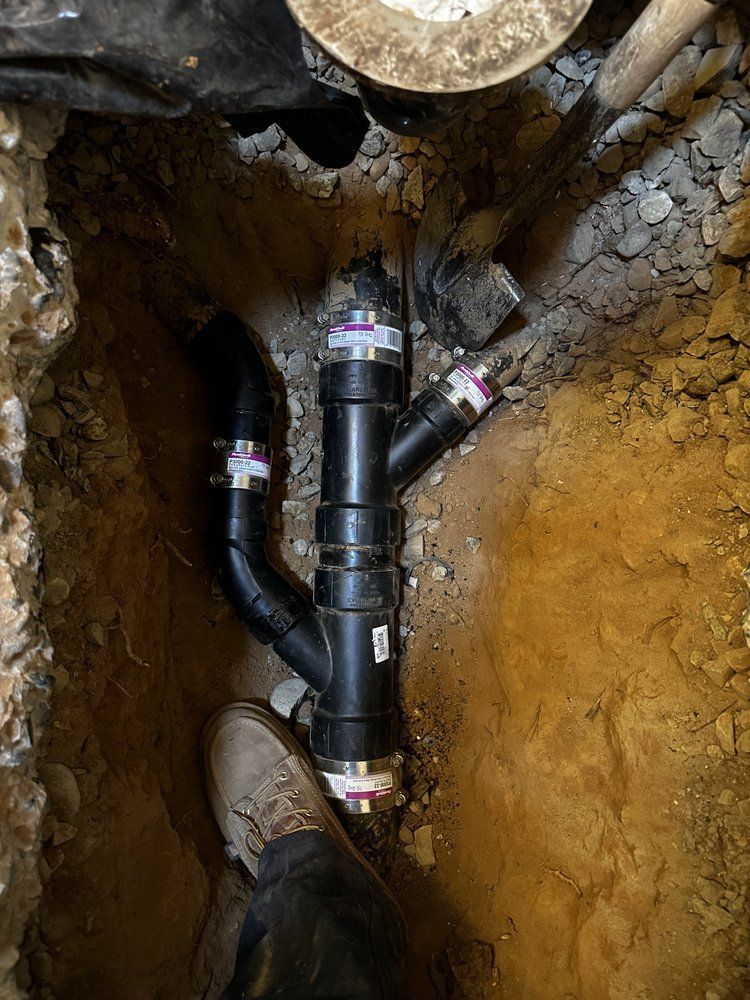 Plumbing pipes joined in a dirt trench, with black PVC fittings and a concrete opening above.