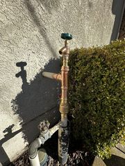 Outdoor copper water pipe and faucet beside a hedge, mounted against a stucco wall.