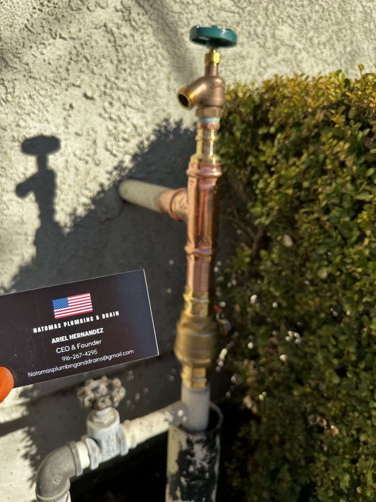 Outdoor brass water pipe with green valve beside a shrub and a small sign on a stucco wall