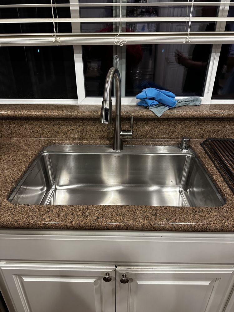 Kitchen sink under a window with blinds, blue gloves on the sill, and granite countertop surrounding it