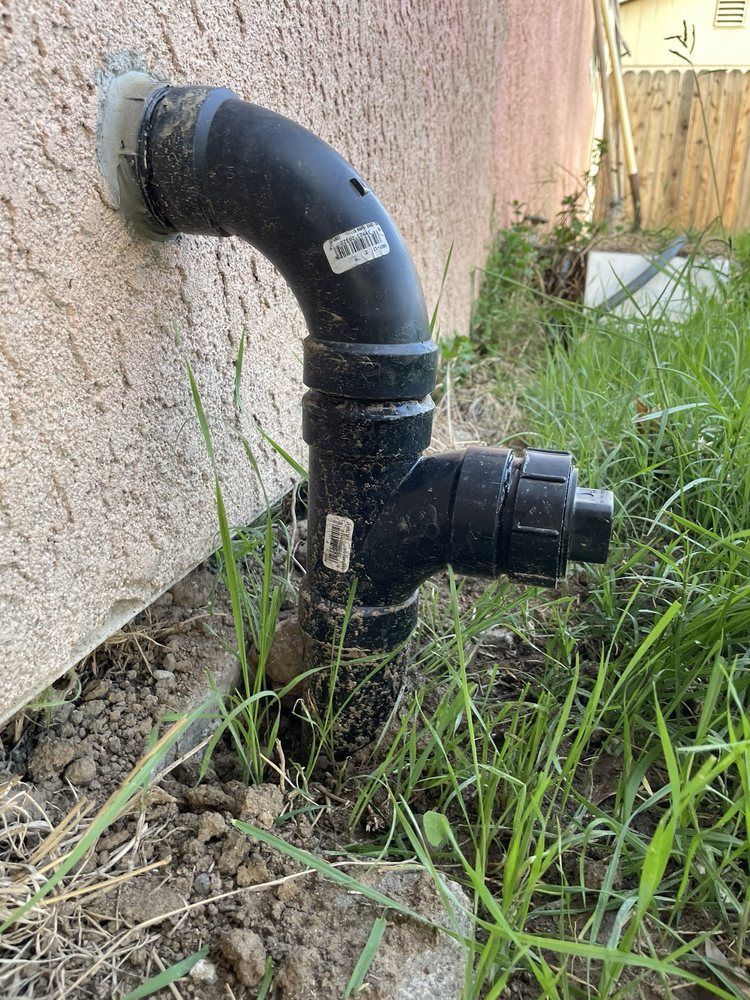 Black outdoor pipe and valve assembly protruding from a stucco wall in grass.