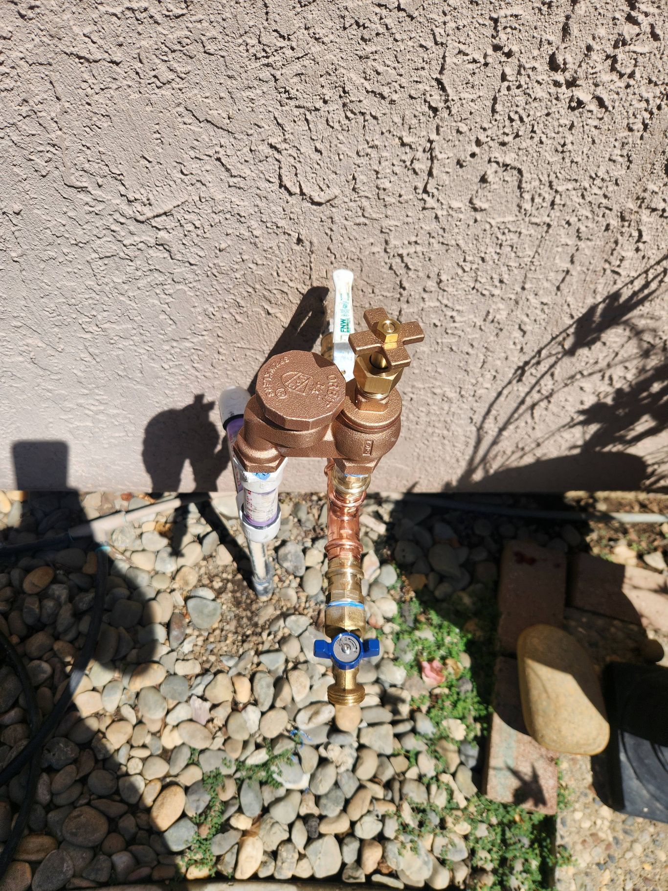 Outdoor water meter and valves beside a stucco wall, surrounded by rocks and gravel.
