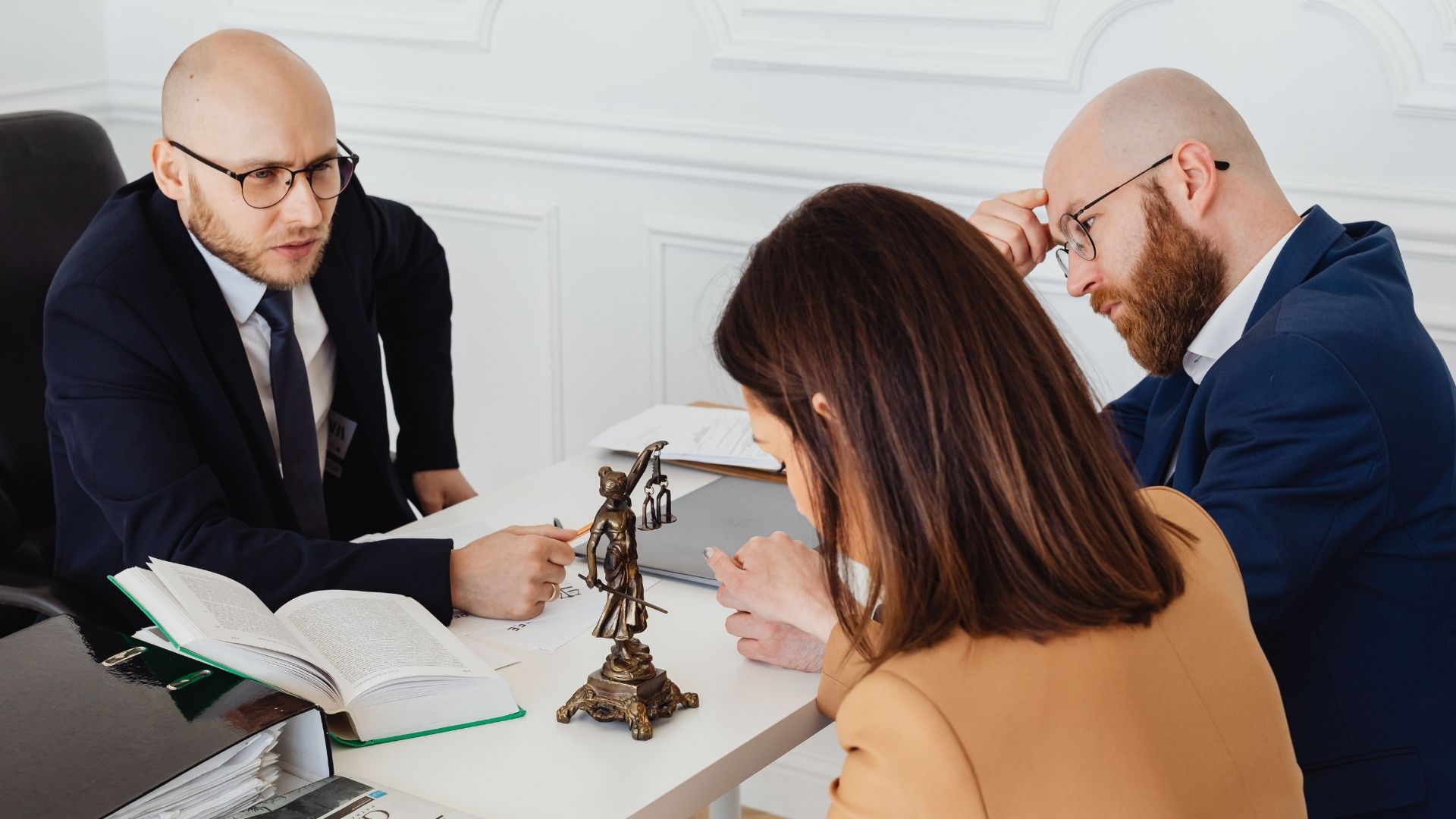 Three people in a meeting reviewing documents across a desk with legal scales centerpiece