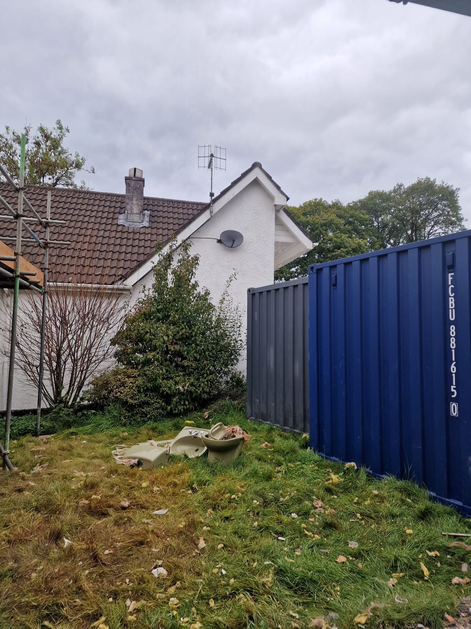 A backyard scene with a white house, blue shipping container, and overgrown grass under an overcast sky.