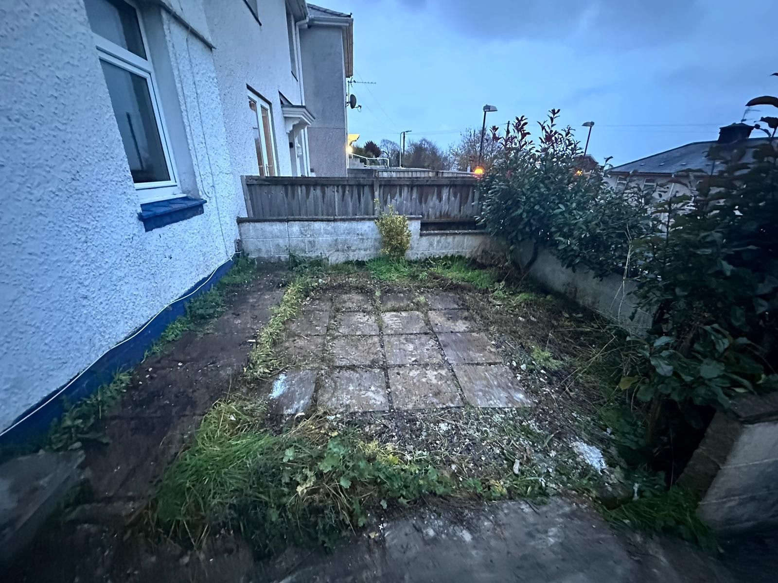 A small, overgrown patio area with concrete paving stones and patchy grass against a building exterior.