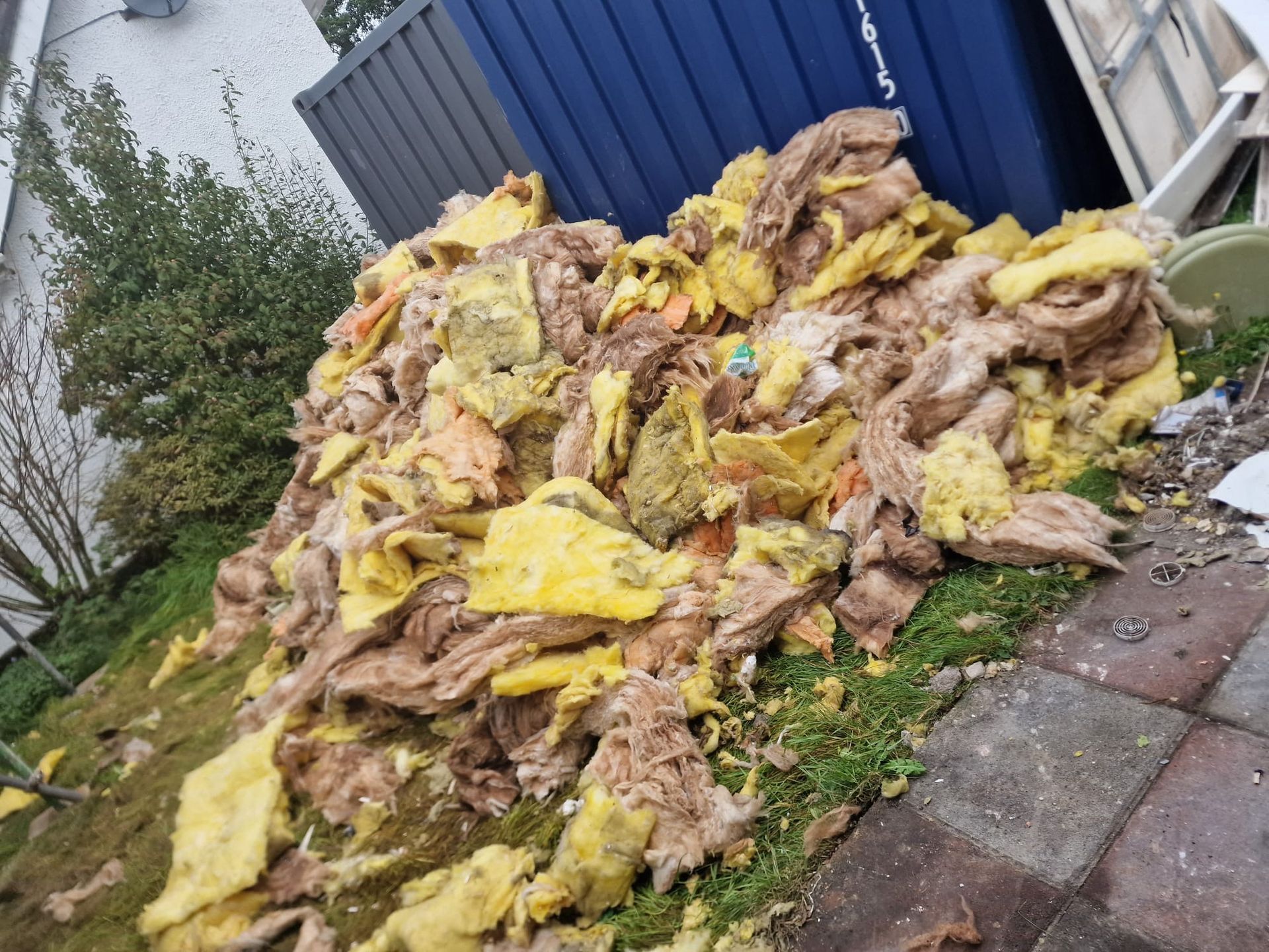 A large pile of yellow and brown insulation debris sits on grass and paving stones outside a building.