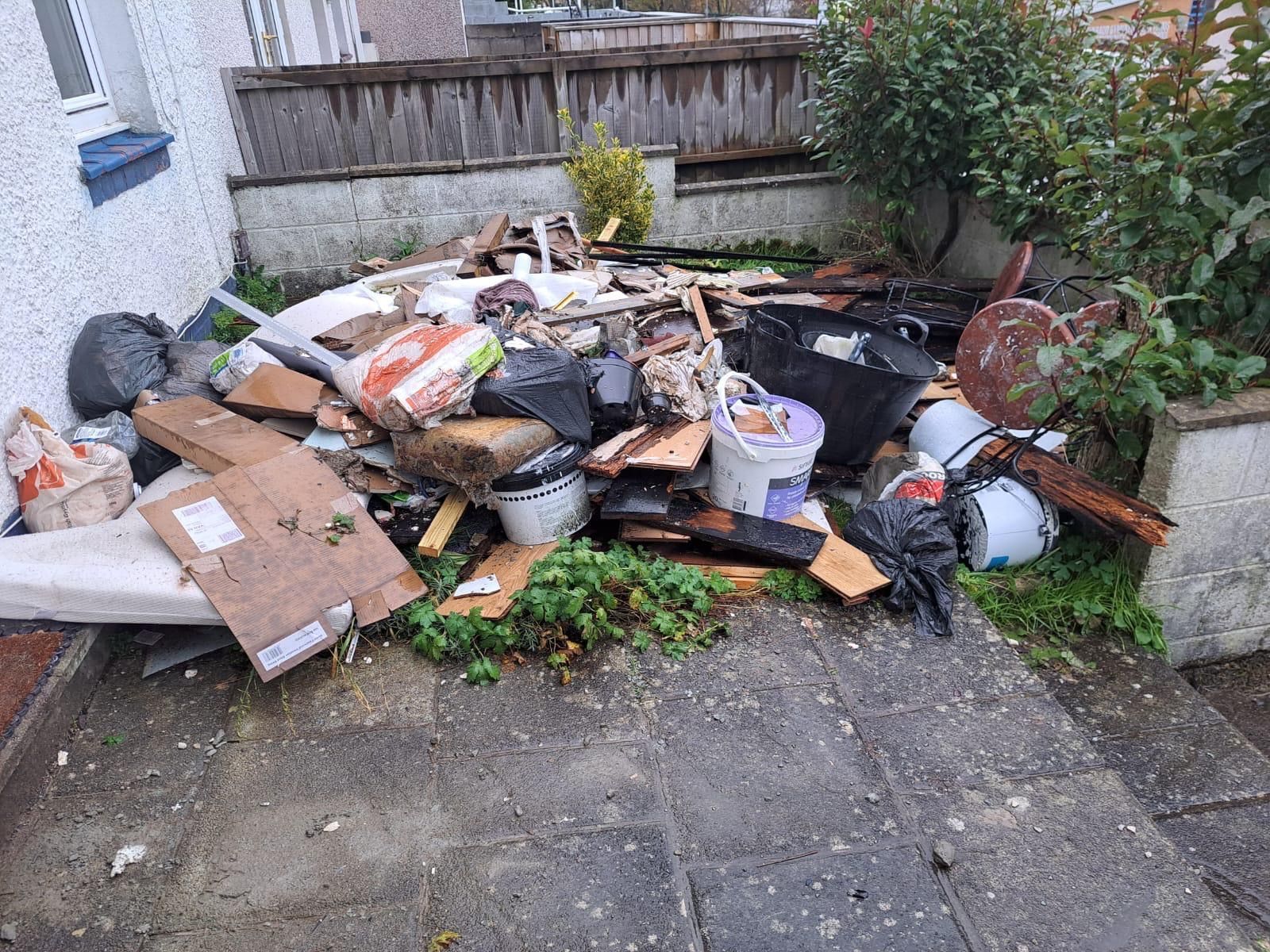 A pile of trash, including wood, paint cans, and bags, on a paved patio outside a house.