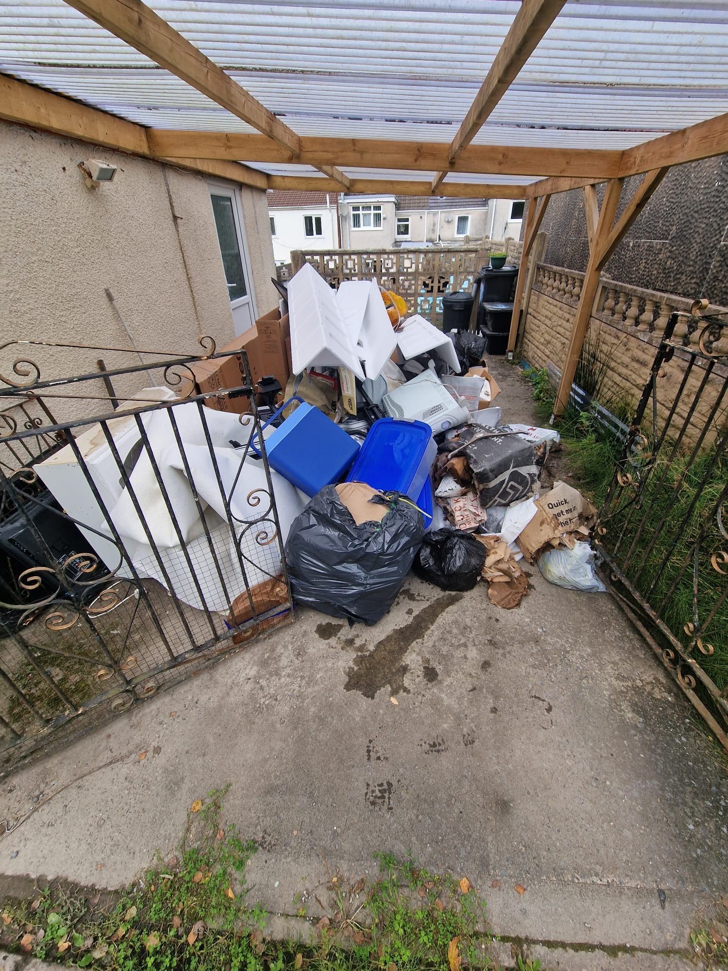 Overfull alleyway with trash, blue bins, and a black trash bag, under a wooden and plastic roof structure.