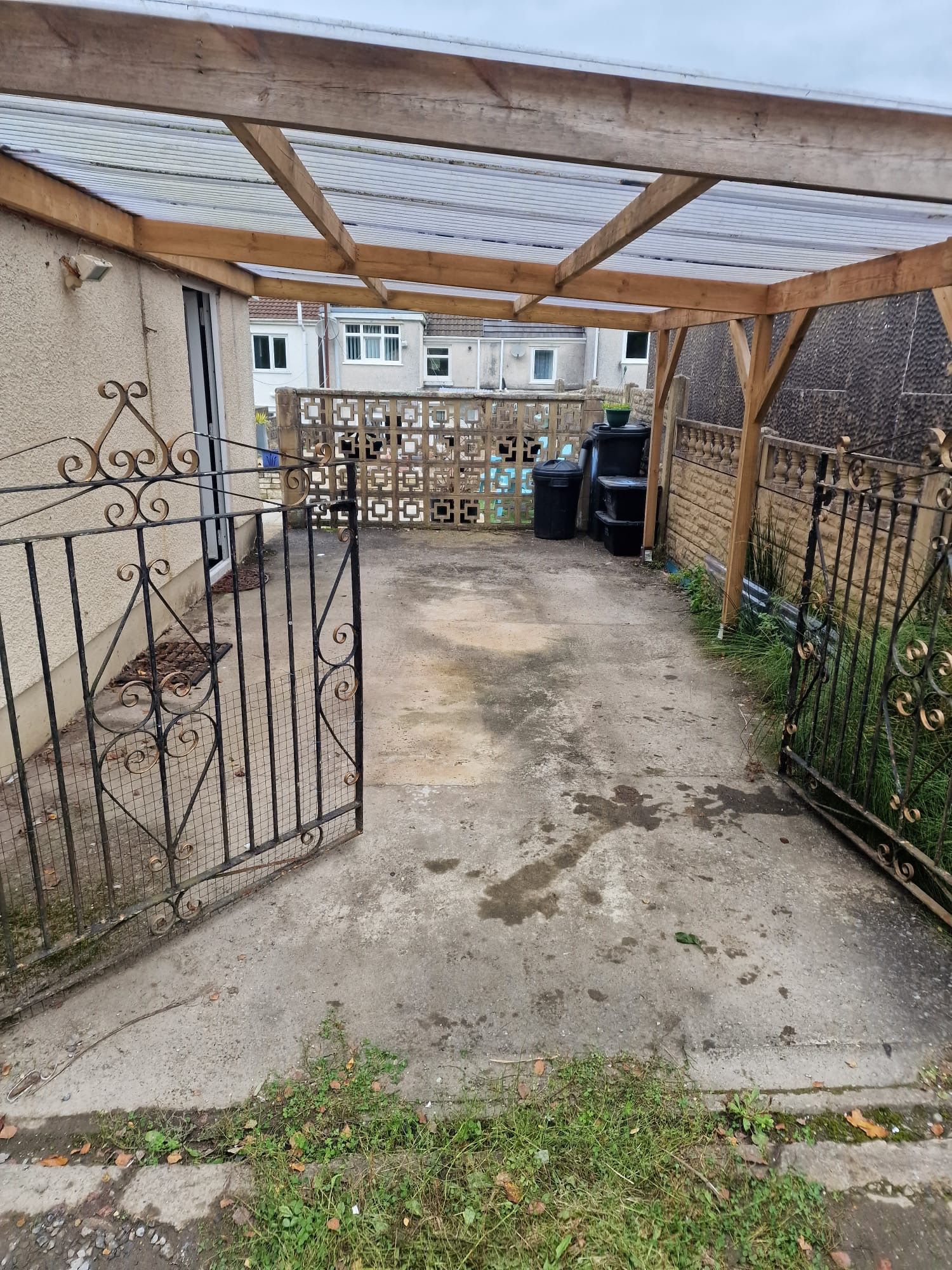 Driveway with an open wrought iron gate, covered by a wooden pergola. The concrete is weathered. Behind the pergola is a fence and buildings.