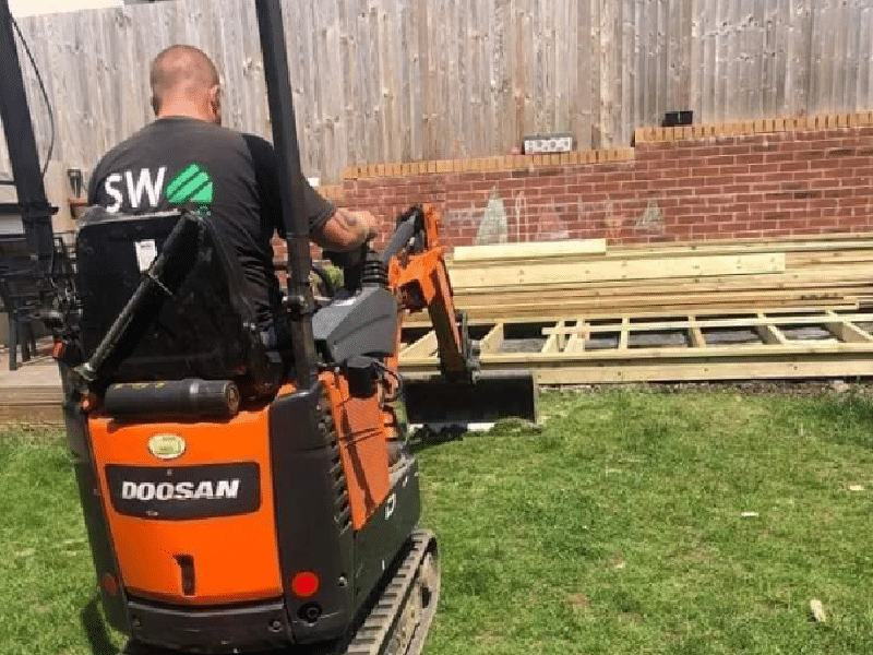 A person operates an orange Doosan excavator on a grassy lawn near wooden materials. Behind the person is a wooden fence and a brick wall.