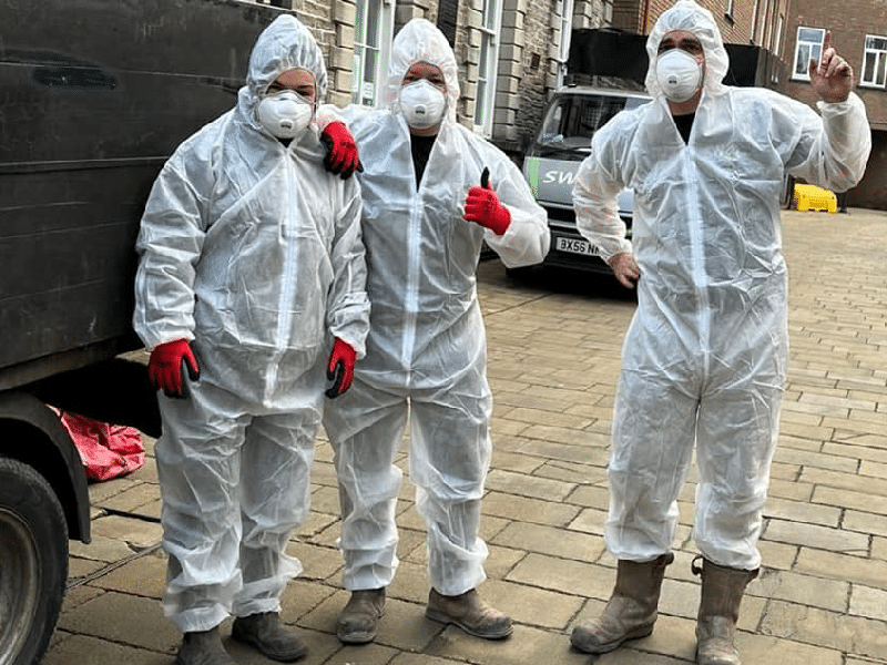 Three people wearing white protective suits, masks, and gloves stand next to a truck. One gives a thumbs-up.