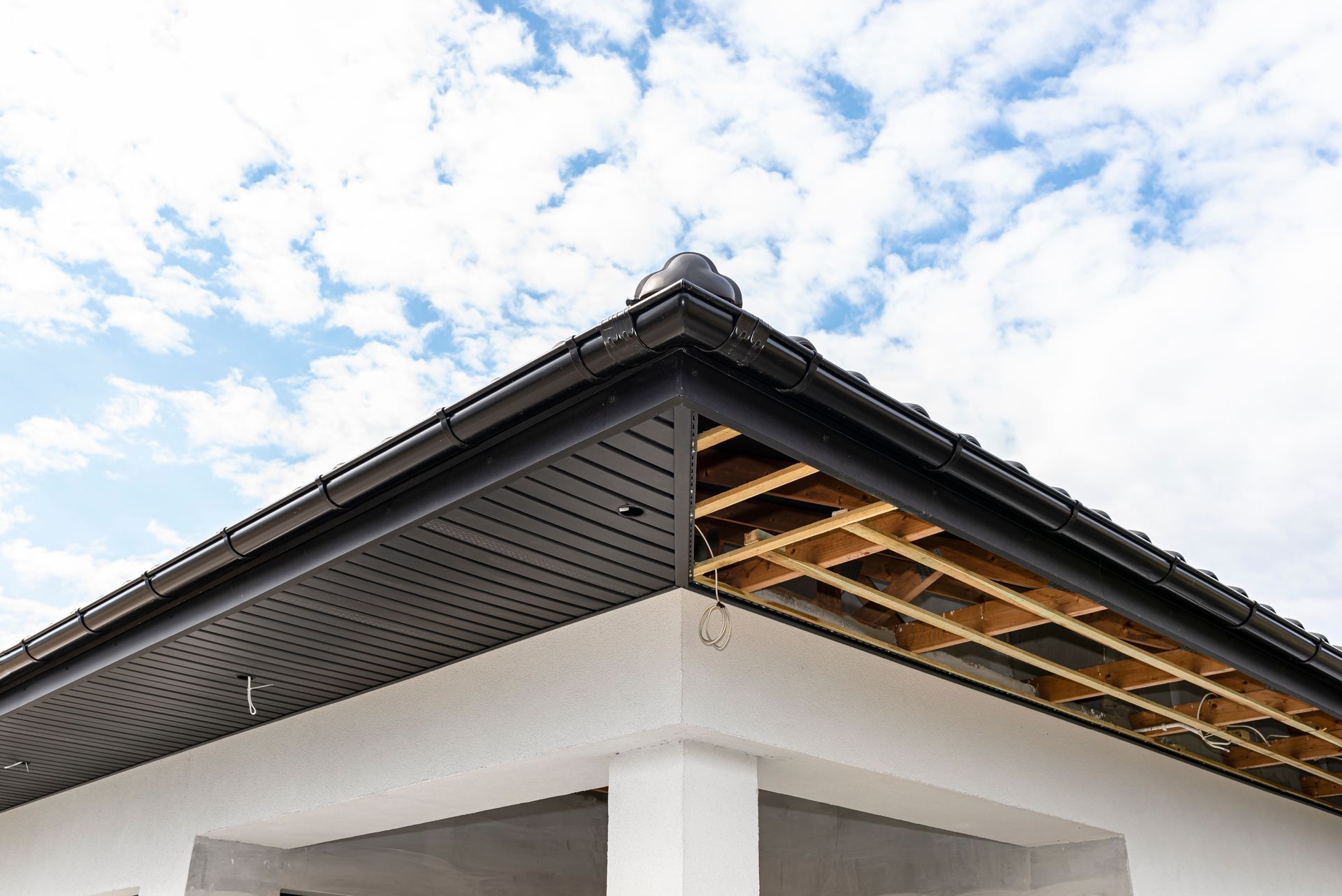 Corner of a building with black roof trim and exposed wooden beams, white exterior against a cloudy sky.