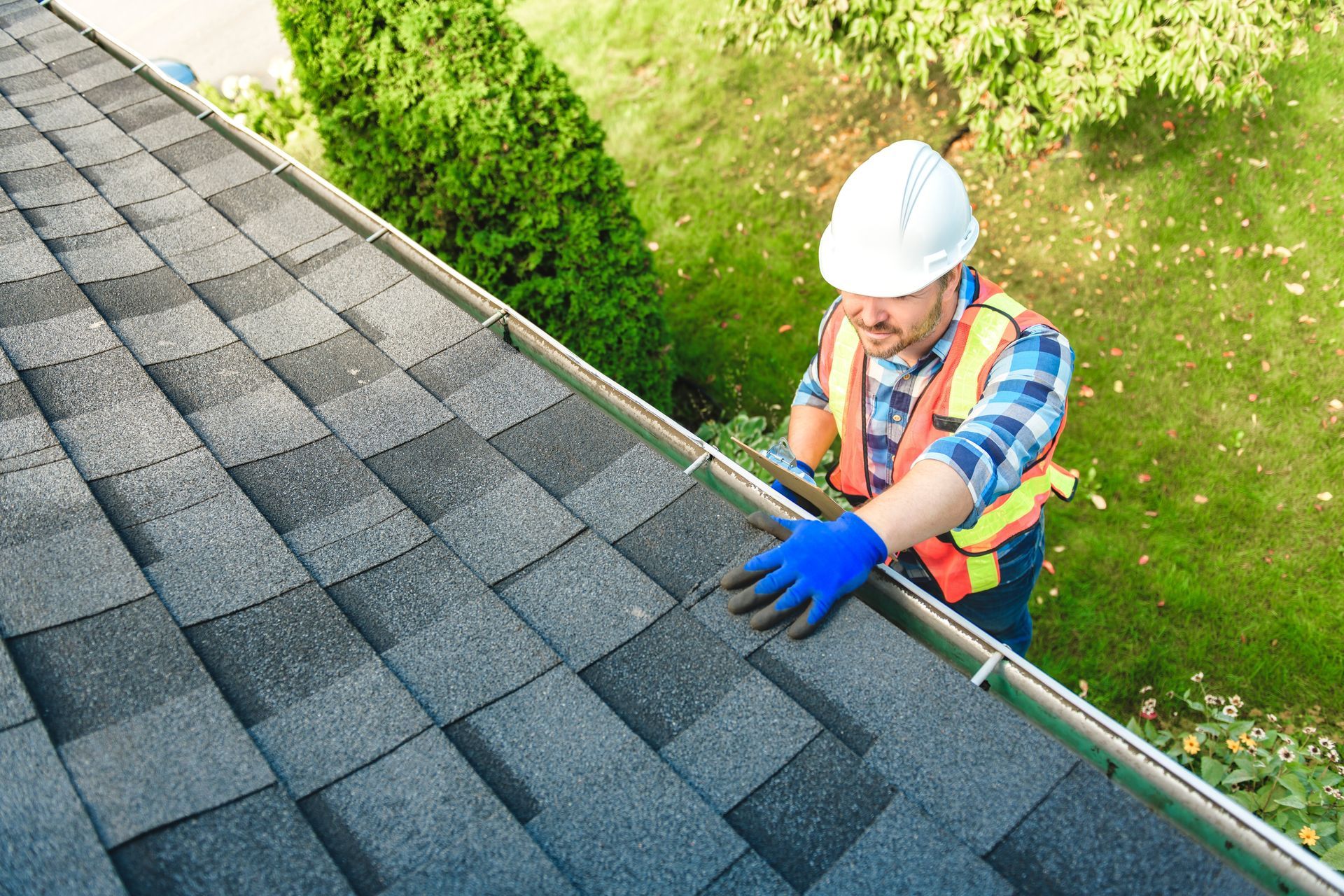 Roofer on a sloped roof wearing a hard hat, inspecting shingles, green foliage in the background.